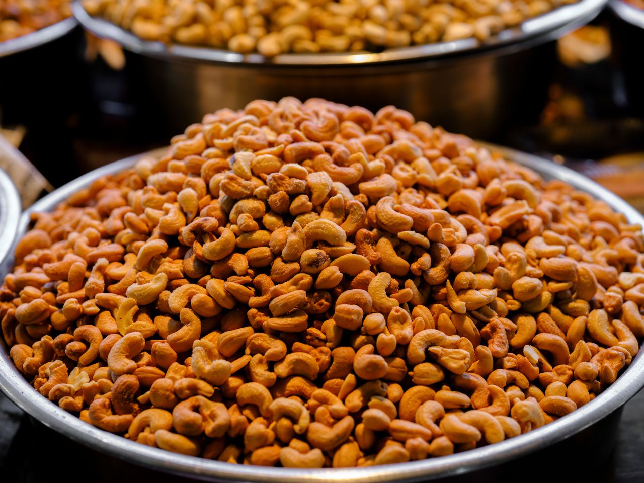 A close-up shot of freshly roasted cashews in a large metal bowl at a bustling market.